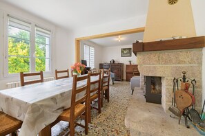 A dining area with a table set for six, a fireplace, and a view of trees through the windows. - Gîte Chez Madeleine: Private Terrace, Garden, and Wi-Fi in Coutances (Coutances)