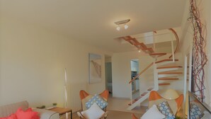 Un salon moderne avec un canapé, des fauteuils, une table basse en verre, un escalier et une cuisine en arrière-plan.