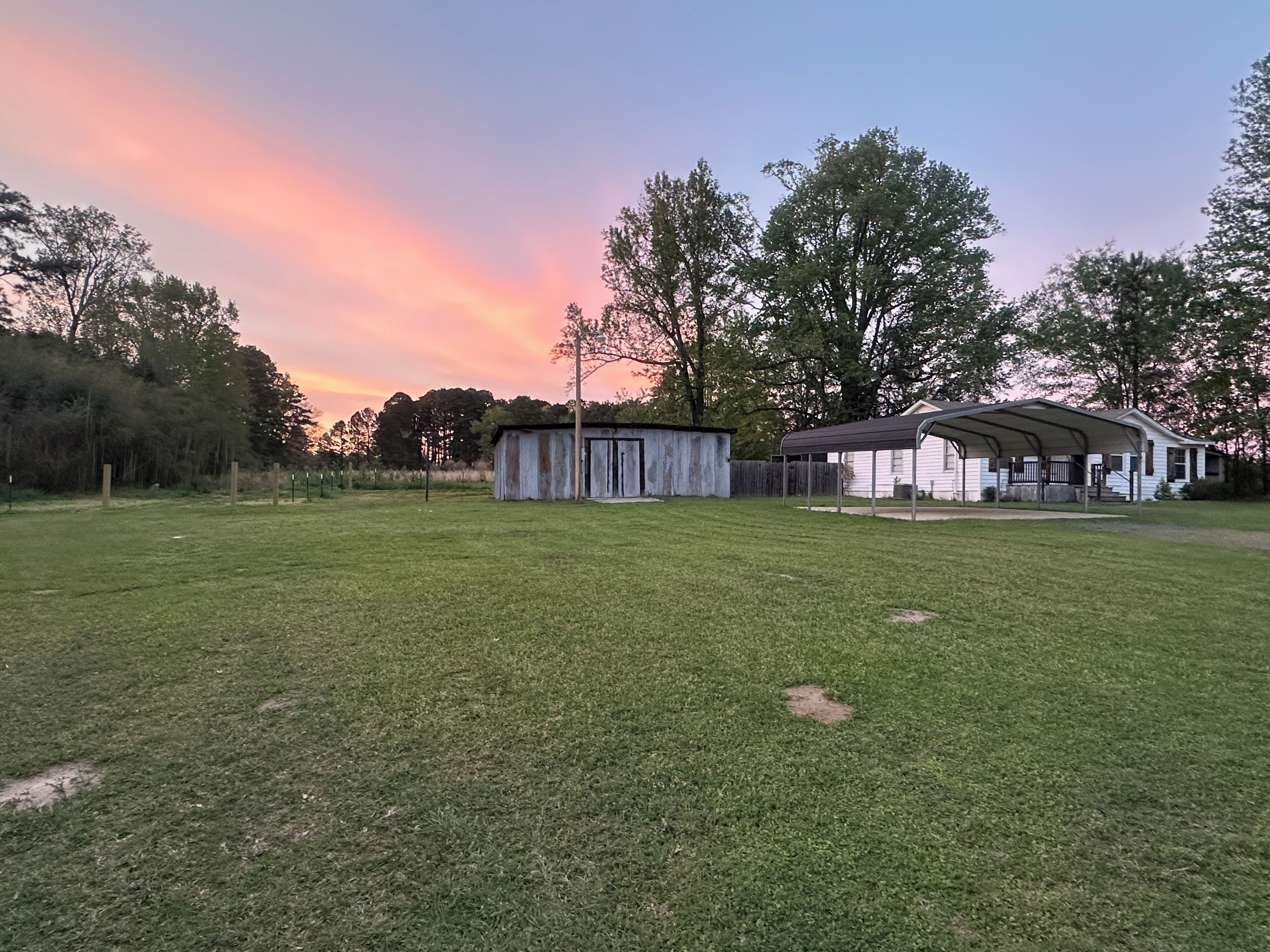 A grassy field with a small shed and a covered structure, surrounded by trees and a house in the background.