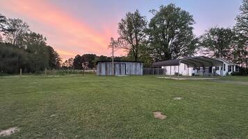 A grassy field with a small shed and a covered structure, surrounded by trees and a house in the background.