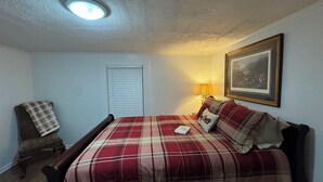 Un dormitorio con cama, una silla, una lámpara, una ventana con persianas y un cuadro en la pared.