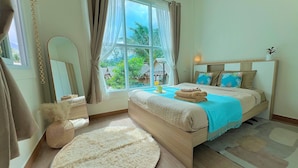 Une chambre avec une grande fenêtre, un lit recouvert d’une couverture bleue, un tapis rond et une tête de lit en bois.