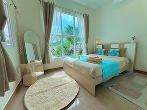 A bedroom with a large window, a bed with a blue blanket, a round rug, and a wooden headboard. - Stylish Tropical Cottage in Chaloklum Heart, 3 Min Walk to Beach (Tambon Ko Pha-ngan)