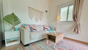 Un salon comprenant un canapé, une table d’appoint, un tapis à motifs, une décoration murale en macramé et une fenêtre avec des rideaux.