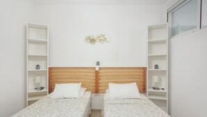 Una habitación pequeña y limpia con dos camas individuales, cabecero de madera y paredes blancas.
