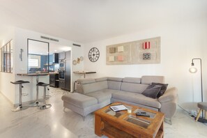A modern living room with a sectional sofa, a glass coffee table, and a kitchen area visible through an open door. - Vacation Home 'Adosado A Un Paso Del Mar' with Shared Pool, Wi-Fi and Air Conditioning (Xàbia)