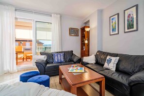 Una sala de estar con un sofá de cuero negro, una mesa de centro de madera y un otoman azul. Se observa una estantería con una lámpara y una ventana con cortinas.