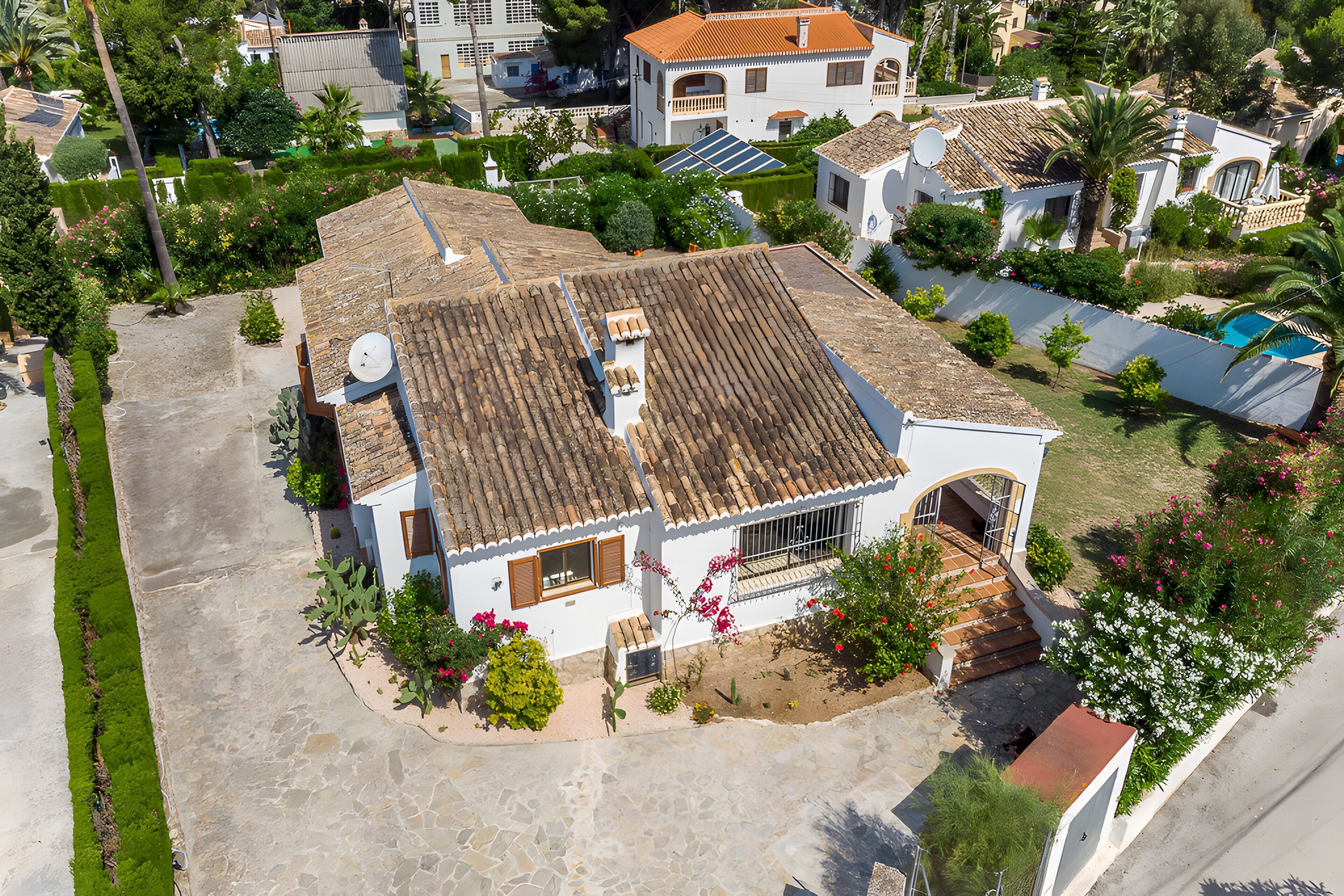 Una casa residencial con techo de tejas, una antena parabólica y un pequeño jardín con plantas en flor.