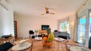Un coin repas avec une table dressée pour quatre personnes, un ventilateur de plafond, un téléviseur et une grande fenêtre.