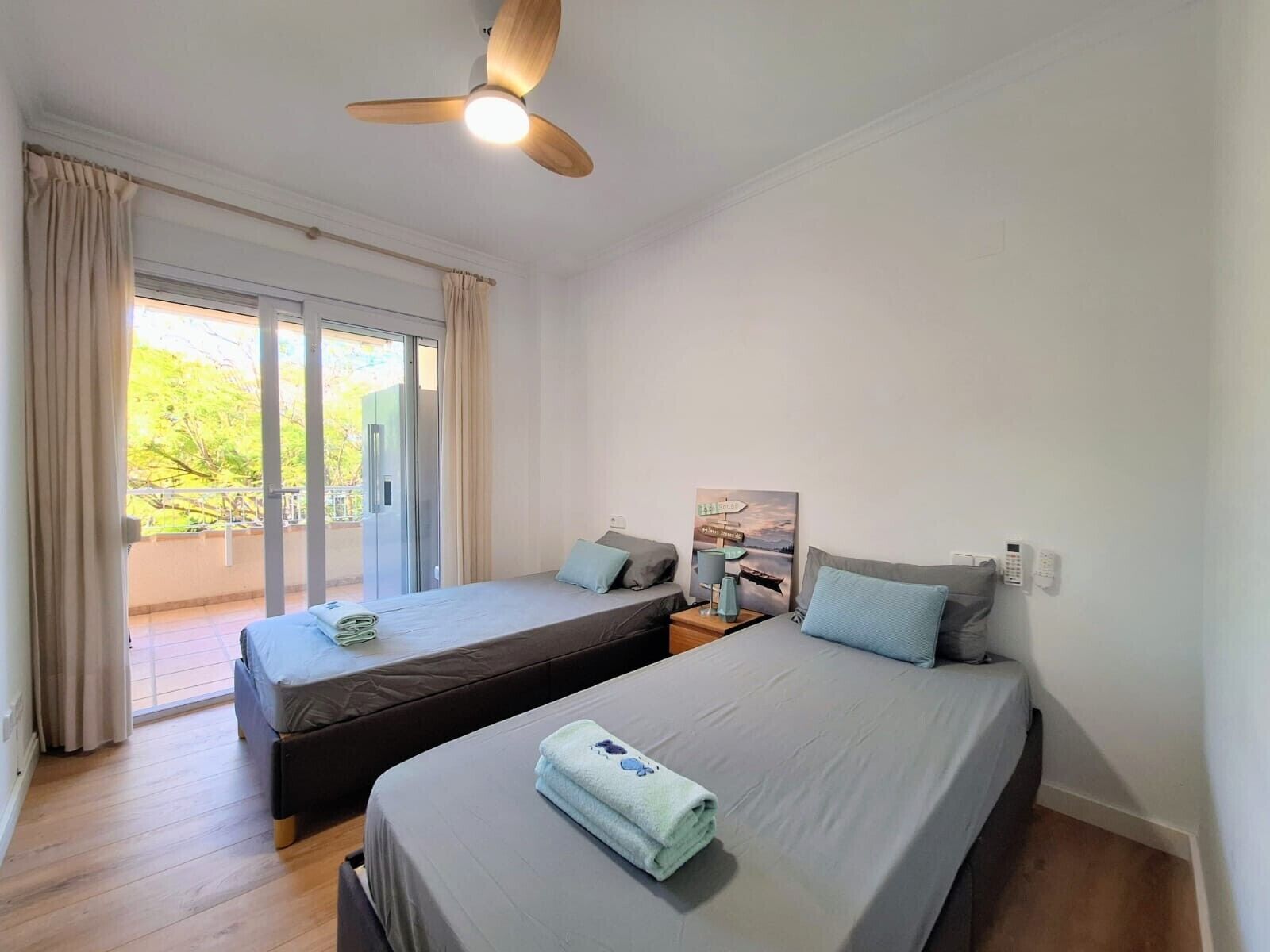 A hotel room with two beds, a ceiling fan, a painting on the wall, and a view through the glass door.