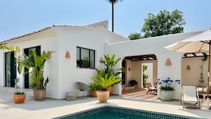 Un área de piscina moderna con piscina de aguas cristalinas, plantas en macetas, mobiliario de exterior y un edificio blanco con tejado de azulejos.