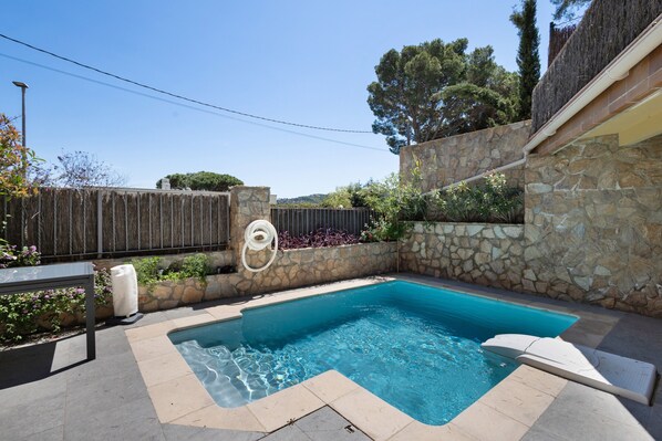 Una piscina con muros de piedra, cerca de madera y una mesa con una silla.