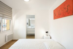 Un dormitorio con cama, ventana, radiador, pared blanca y baño visible a través de una puerta abierta.