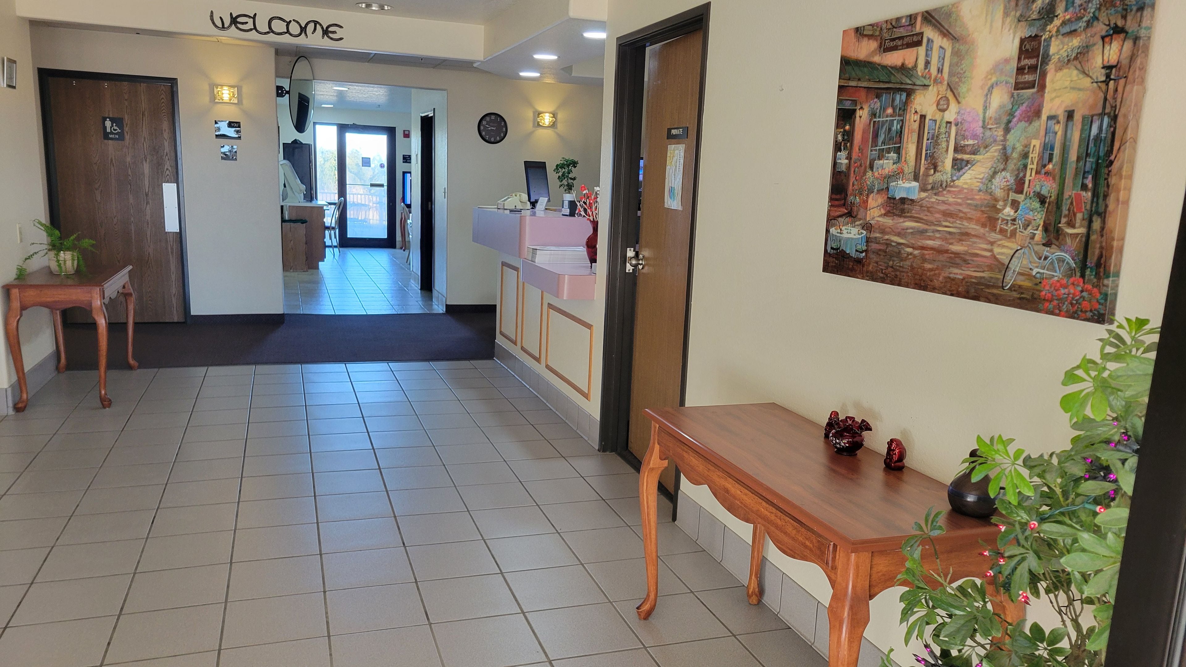 A hotel reception area with a reception desk, a 'Welcome' sign, a clock, and a painting on the wall.