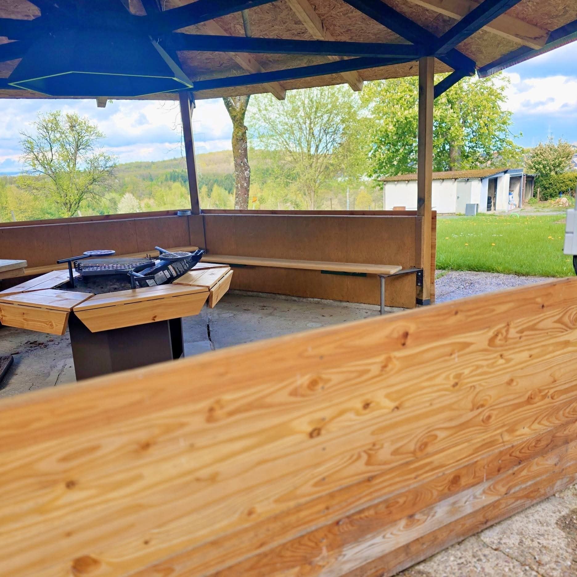 En utomhusplats med en träbänk och en grill, placerad under en överbyggnad, med utsikt över en gräsyta och träd.