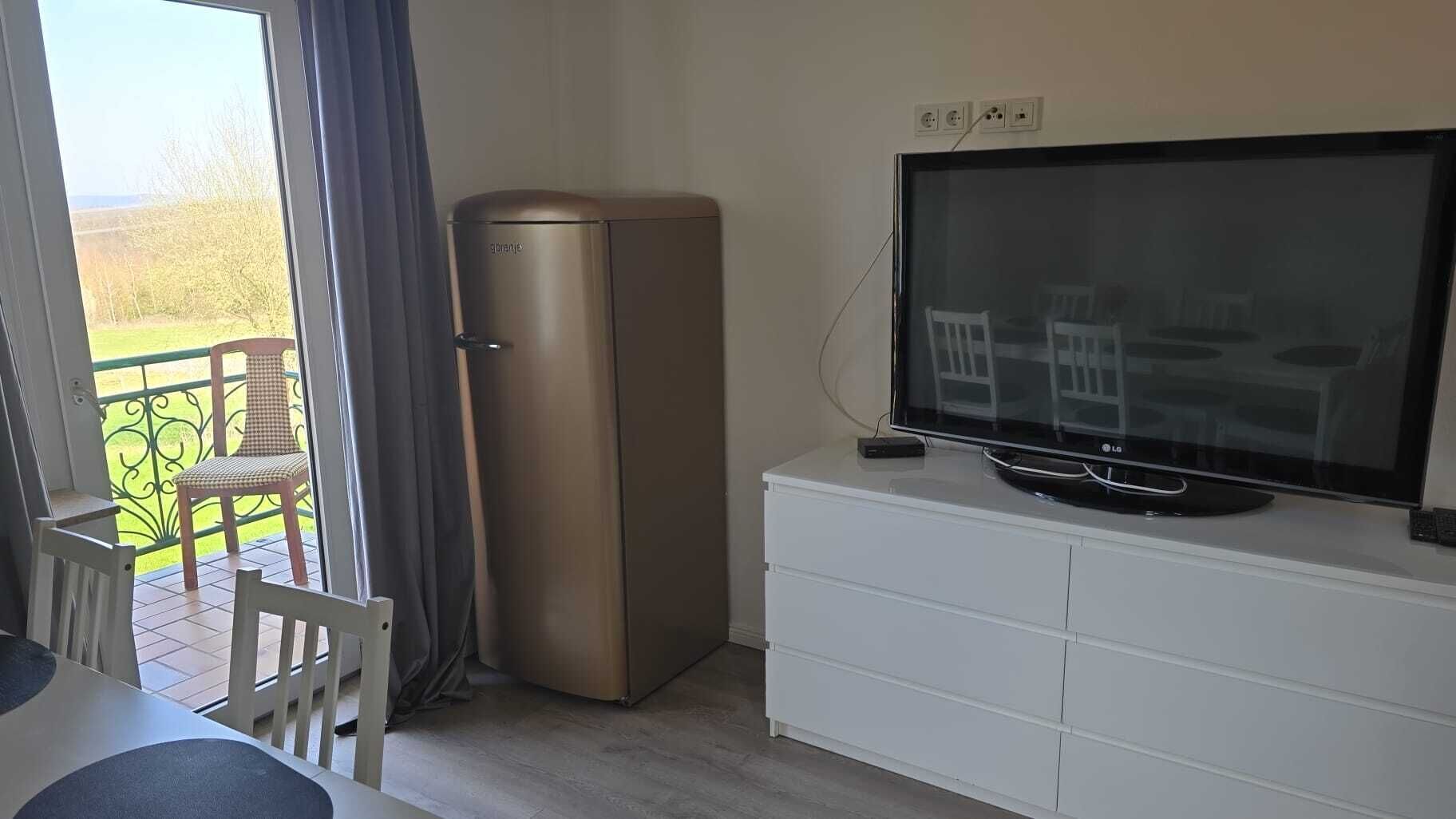 Una habitación con televisor de pantalla plana sobre un mueble blanco, un refrigerador pequeño, una mesa de comedor con sillas y un balcón con una silla.