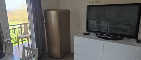 Una habitación con televisor de pantalla plana sobre un mueble blanco, un refrigerador pequeño, una mesa de comedor con sillas y un balcón con una silla.