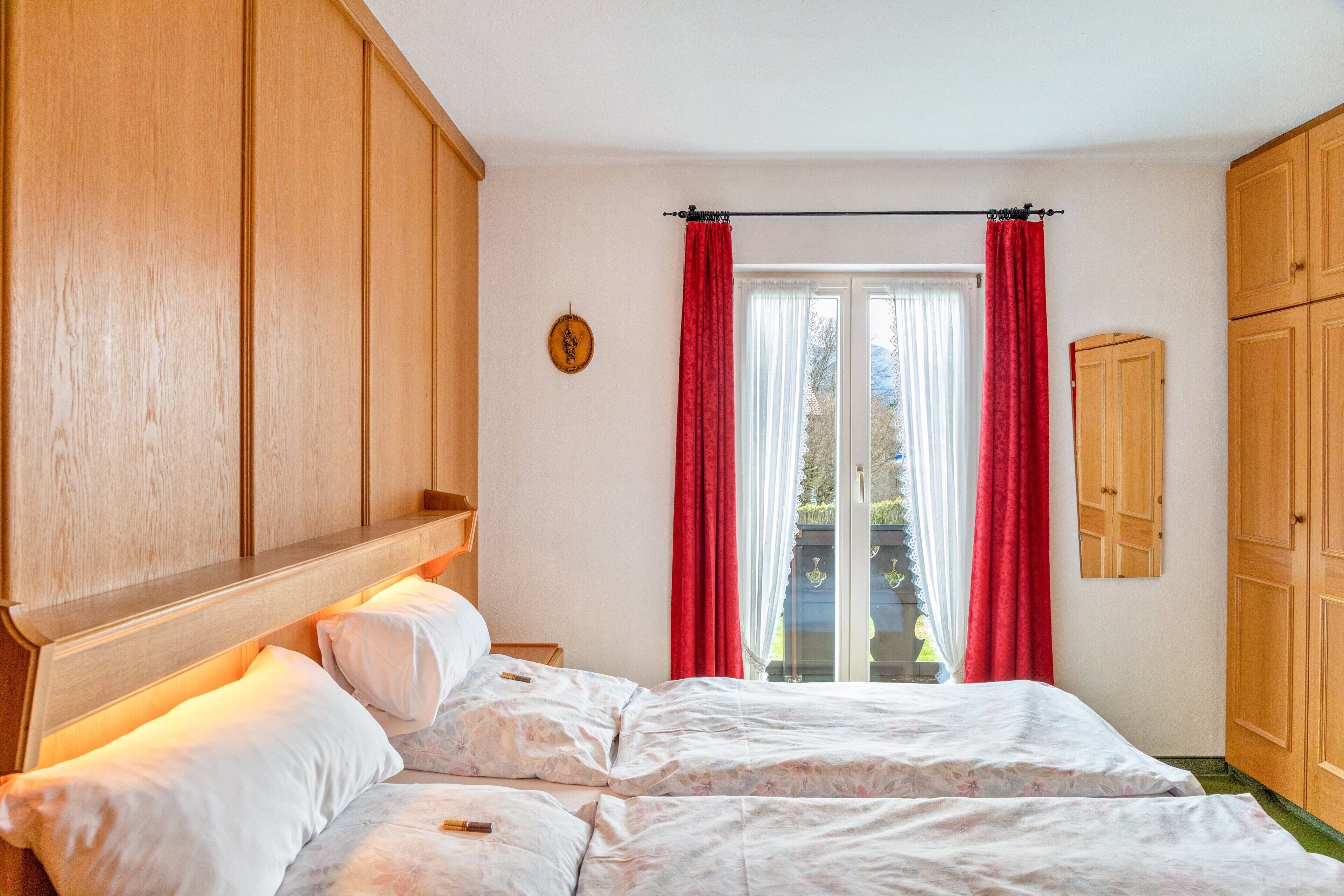 Ein Schlafzimmer mit einem hölzernen Kopfende, einem Bett mit weißer Bettwäsche, einem Fenster mit roten Vorhängen und einem Holzschrank.