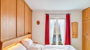 Ein Schlafzimmer mit einem hölzernen Kopfende, einem Bett mit weißer Bettwäsche, einem Fenster mit roten Vorhängen und einem Holzschrank.