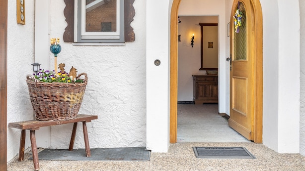A doorway leading to a room with a wooden door, a wicker basket with flowers on a bench, a wall clock, and a framed picture. - Apartment 'Parterre' with Mountain View, Private Terrace and Wi-Fi (Wallgau)