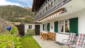 A traditional chalet with a wooden balcony, a small patio with a bench and chairs, and a view of a forested hillside. - Apartment 'Parterre' with Mountain View, Private Terrace and Wi-Fi (Wallgau)