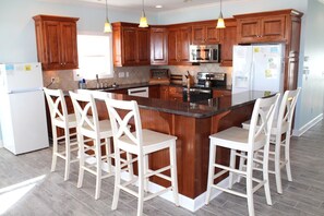 A kitchen with wooden cabinets, a central island with bar stools, a refrigerator, and a microwave. - 7 Bedroom Across the Street from Beach / Access to Oceanfront Pool! (Ocean Isle Beach)