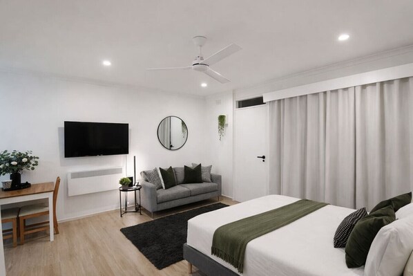 Una camera da letto moderna con un letto, una scrivania, una televisione, una ventola a soffitto e una finestra con tende.