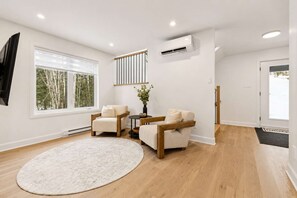 Un salon moderne doté d’une grande fenêtre, d’un tapis rond, de deux fauteuils, d’une petite table et d’un appareil de climatisation.