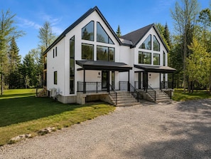 Une maison moderne à deux étages, dotée de grandes fenêtres, d’un porche couvert et d’une allée gravillonnée.