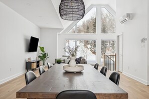 Ein moderner Essraum mit einem langen Holztisch, schwarzen Stühlen, einer weißen Vase in der Mitte und einem großen Fenster mit Blick auf Bäume.