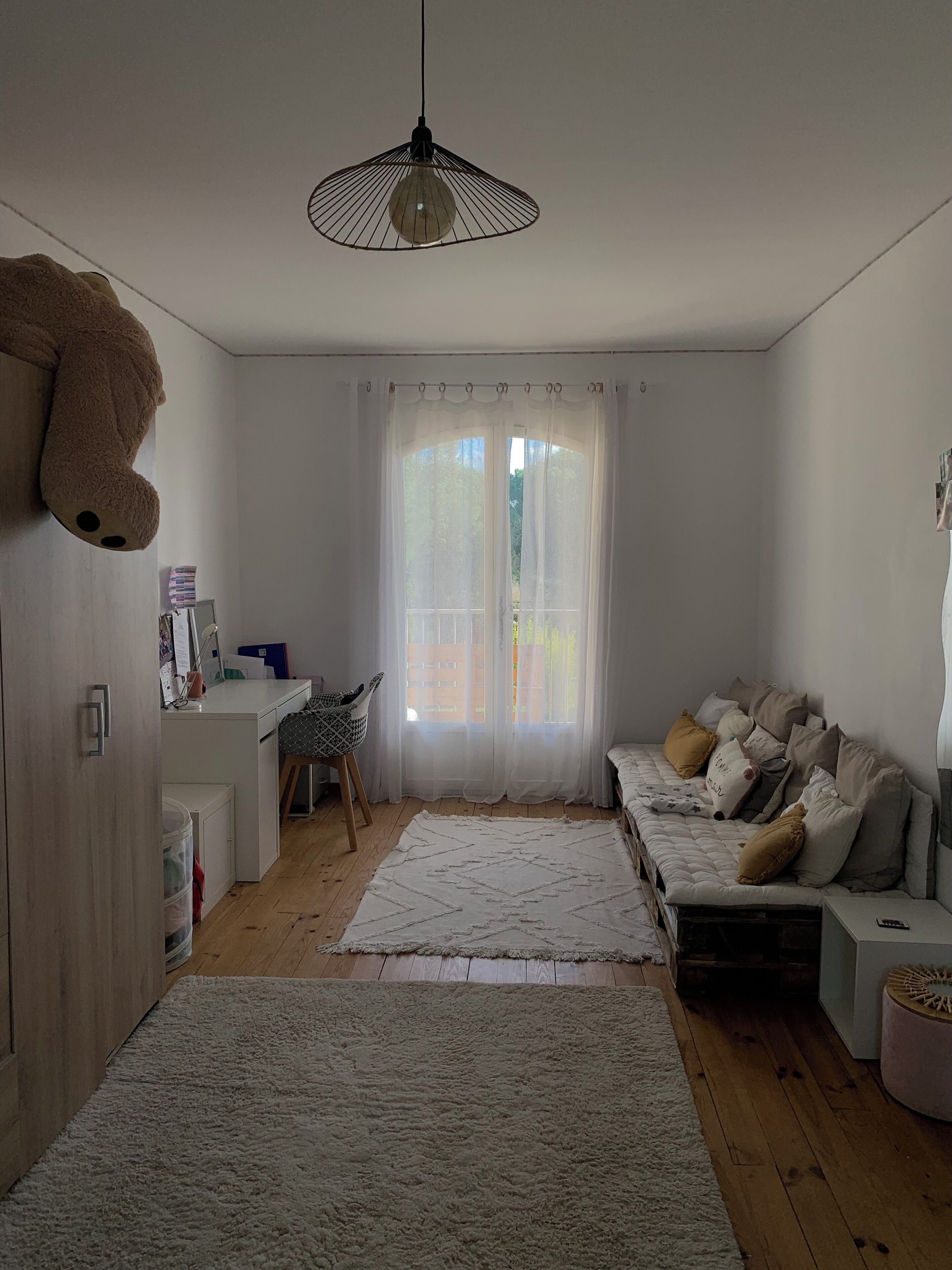 Huoneessa on sänky, työpöytä, tuoli, suuri pehmolelu ja riippuvalaisin.