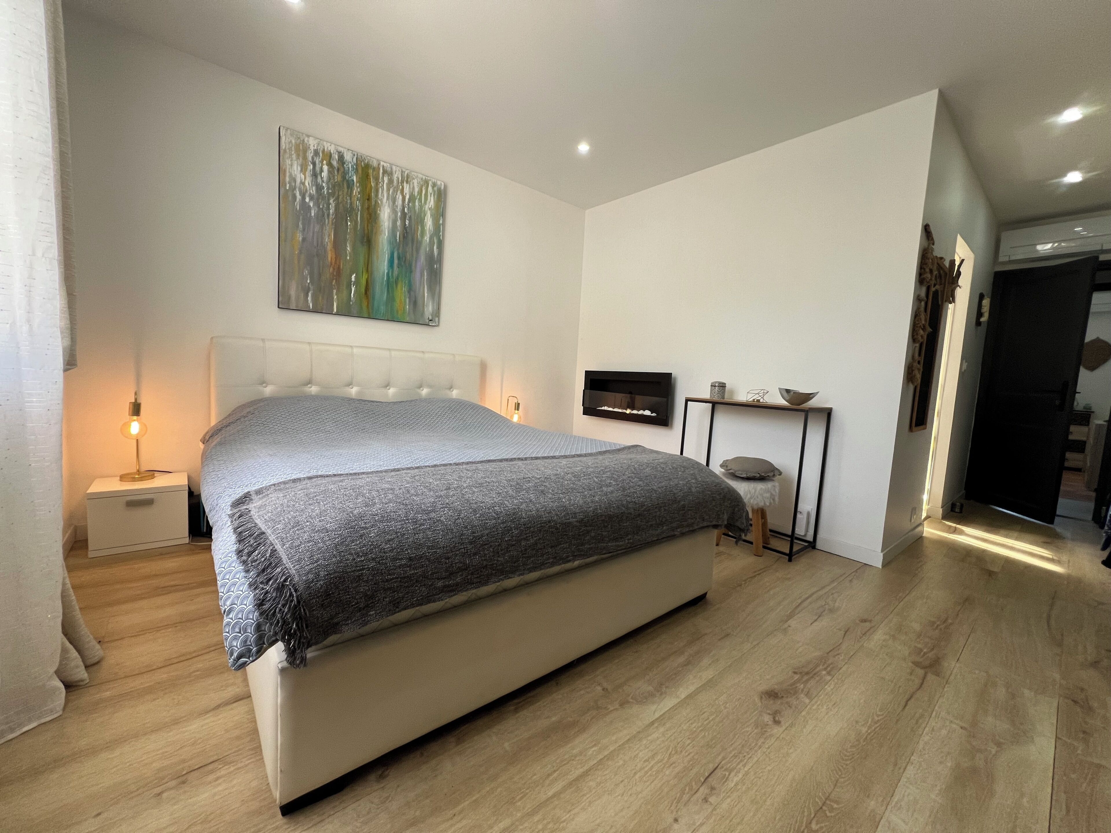 A bedroom with a bed, bedside tables, a wall-mounted shelf, and a door leading to another room.