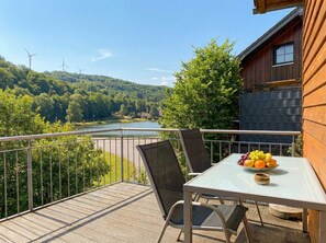 A balcony with a table and chairs, overlooking a river and wind turbines.