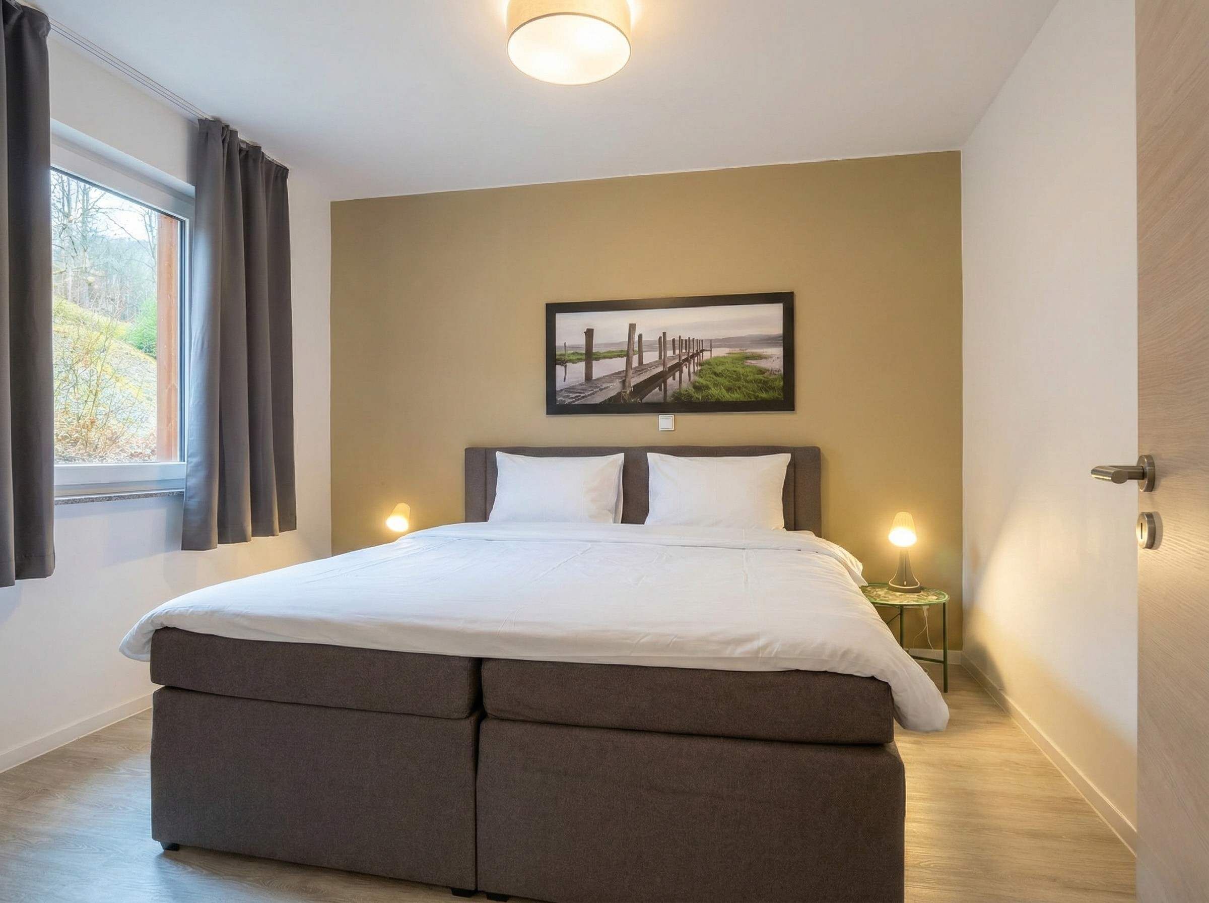 Une chambre à coucher comprenant un lit, deux tables de chevet, une fenêtre avec des rideaux, un tableau accroché au mur et une porte.