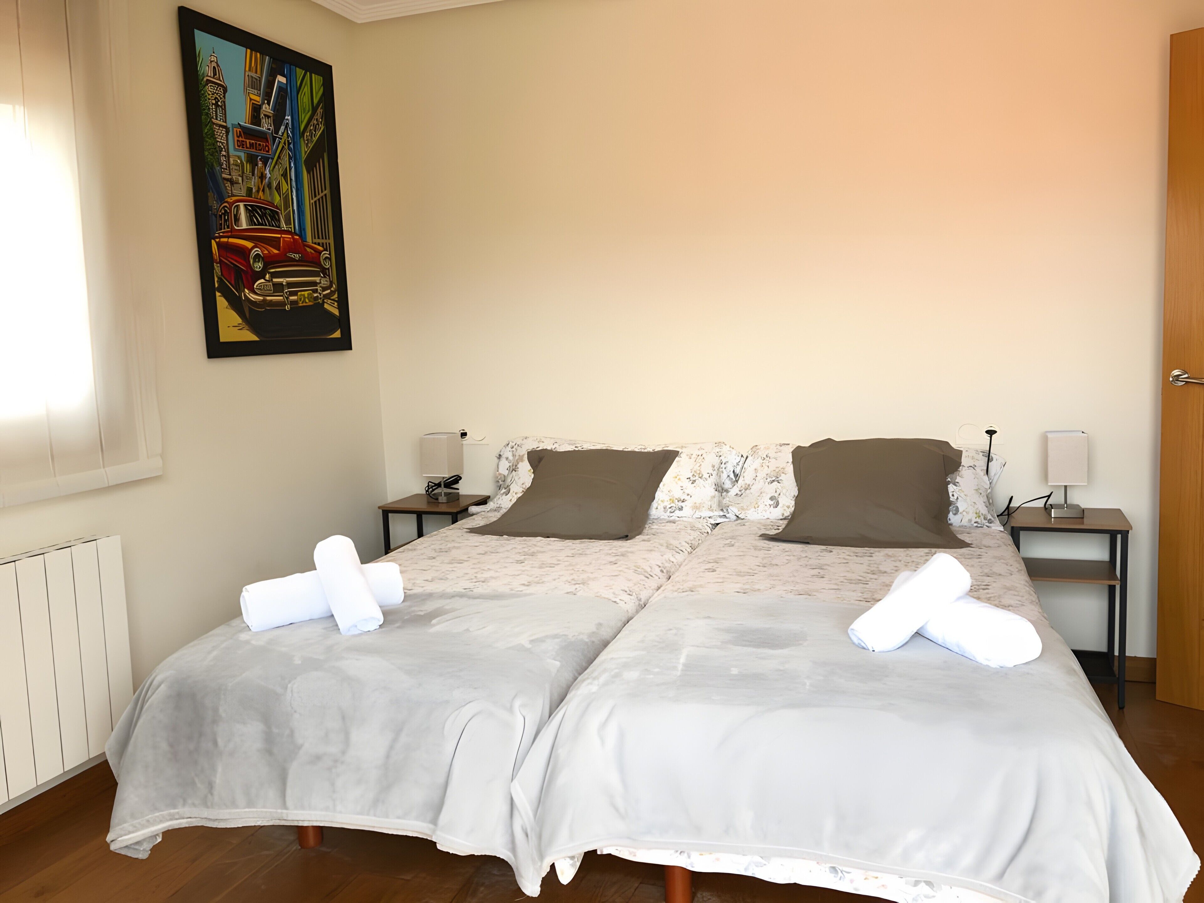 Una cama doble con una colcha gris, dos almohadas marrones, dos toallas blancas, una mesita de noche con una lámpara y un cuadro en la pared.