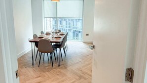 A modern dining area with a wooden herringbone floor, a table set for four, and a large window with a city view. - Anaconda Cut - Campus Accommodation (Salford)