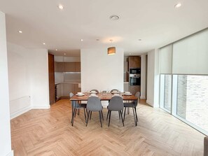 A modern dining area with a wooden herringbone floor, a central table set for four, and a kitchen in the background. - Anaconda Cut - Campus Accommodation (Salford)