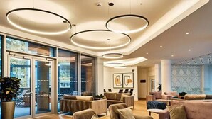 A modern lobby with a glass wall, multiple seating areas, and a ceiling with circular light fixtures. - Anaconda Cut - Campus Accommodation (Salford)