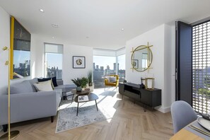 A modern living room with a sofa, coffee table, and a city view through large windows. - Anaconda Cut - Campus Accommodation (Salford)