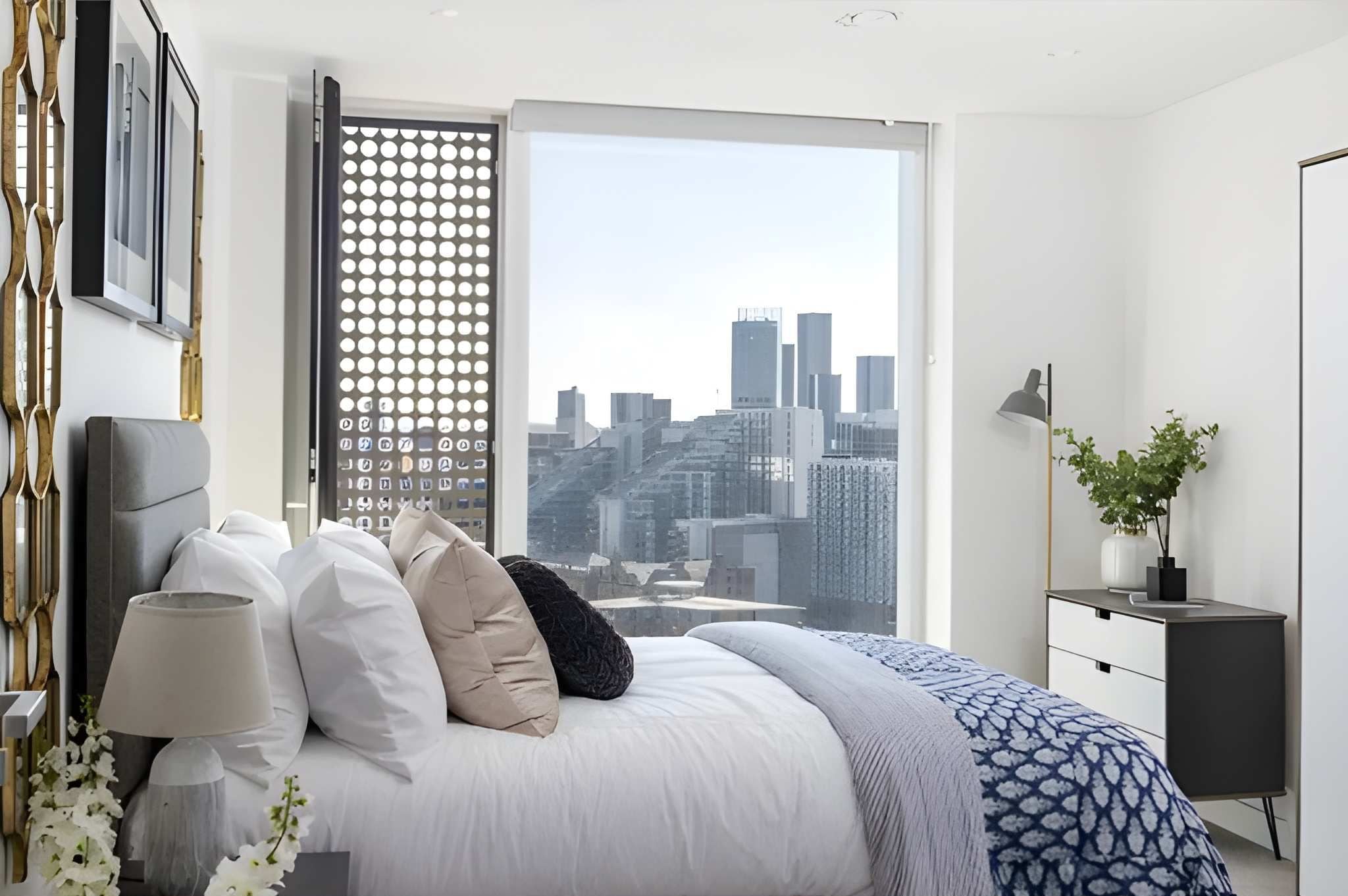 A modern bedroom with a city view, a bed with multiple pillows, a bedside table, and a lamp.