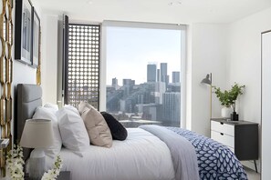 A modern bedroom with a city view, a bed with multiple pillows, a bedside table, and a lamp. - Anaconda Cut - Campus Accommodation (Salford)