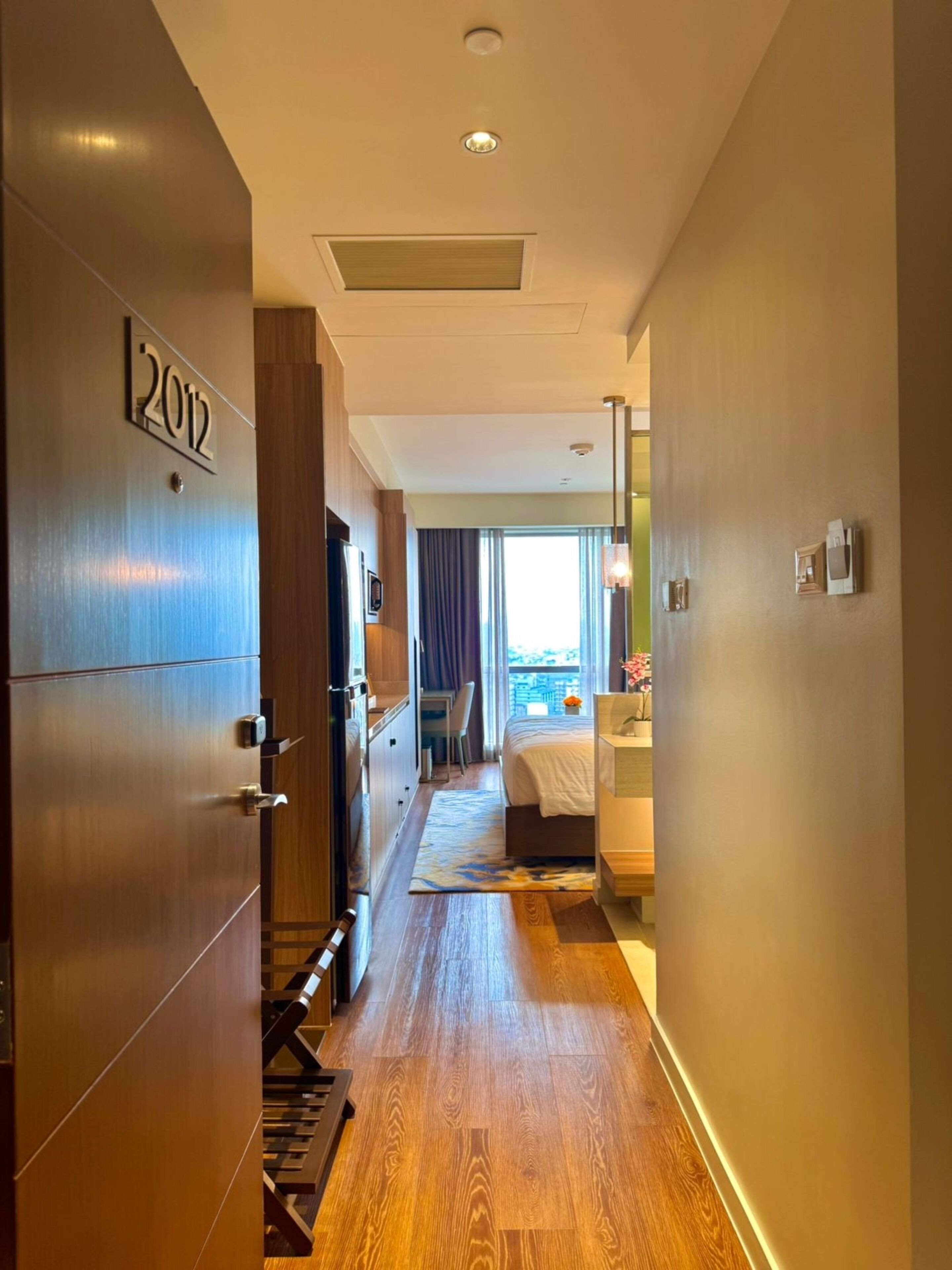 A hotel corridor with a wooden floor, a bed with a white bedspread, a desk with a chair, and a window with curtains.