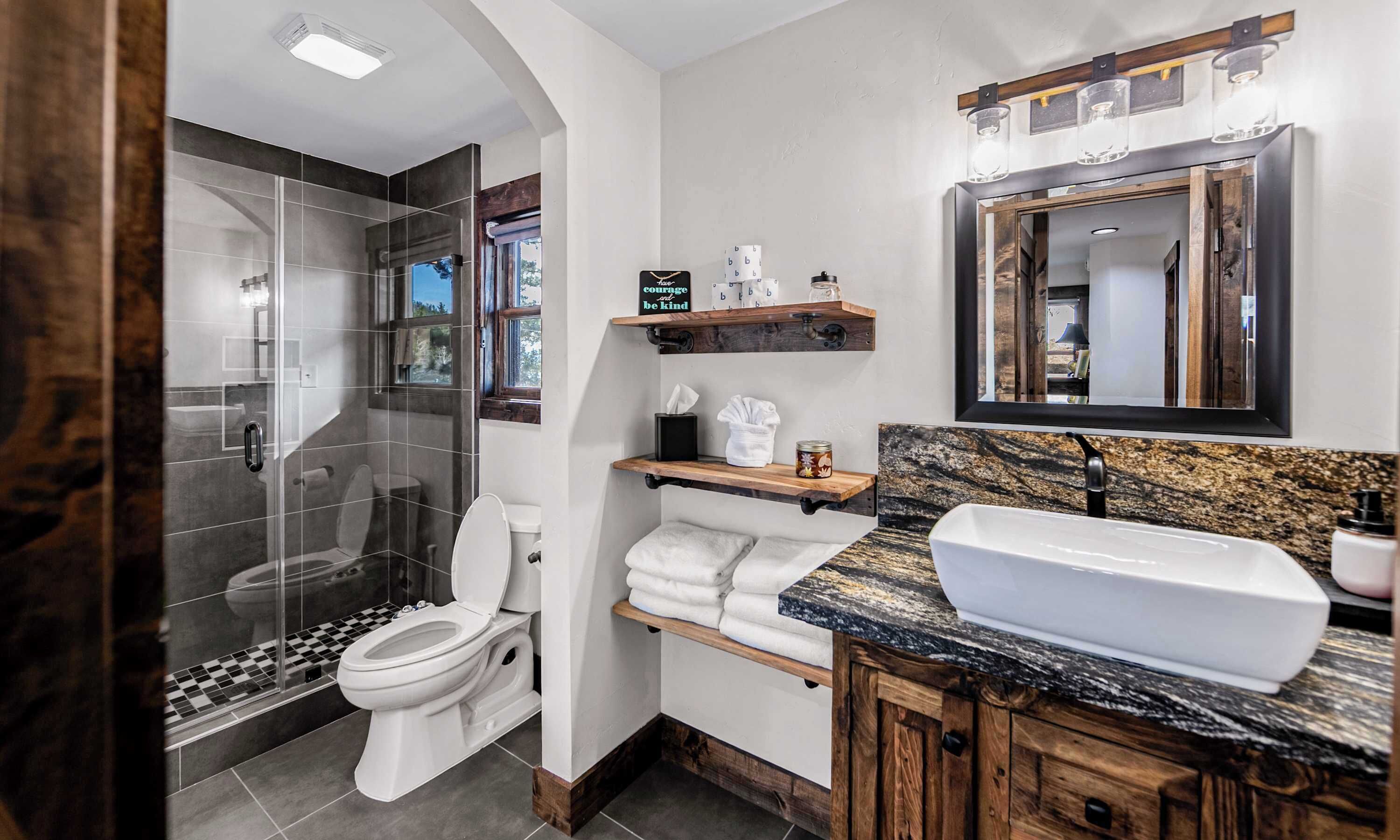 A modern bathroom with a glass shower enclosure, a toilet, a sink with a marble countertop, and wooden shelves with toiletries.