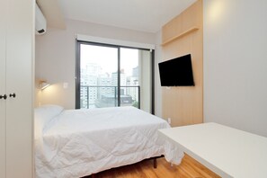 A bedroom with a bed, a bedside table, a TV mounted on the wall, and a large window with a city view. - Blend (São Paulo)