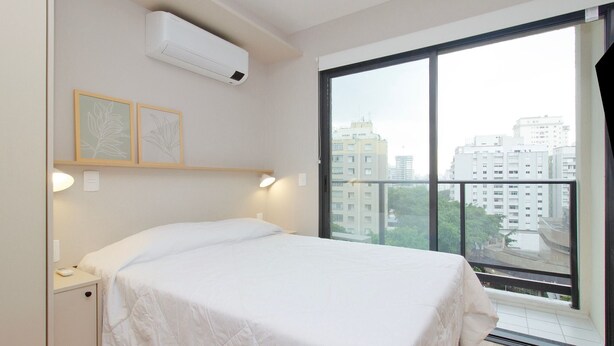 A bedroom with a bed, an air conditioner, a window with a view of buildings, and two framed pictures on the wall. - Blend (São Paulo)