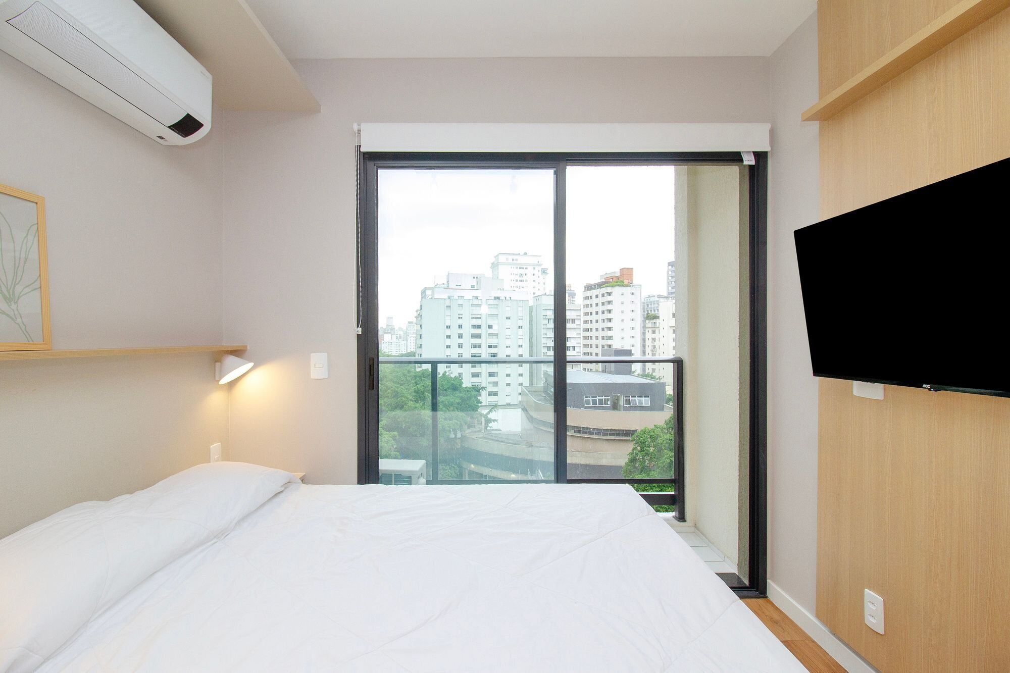 Habitación de hotel con una cama, televisor montado en la pared y un gran ventanal con vista a edificios.