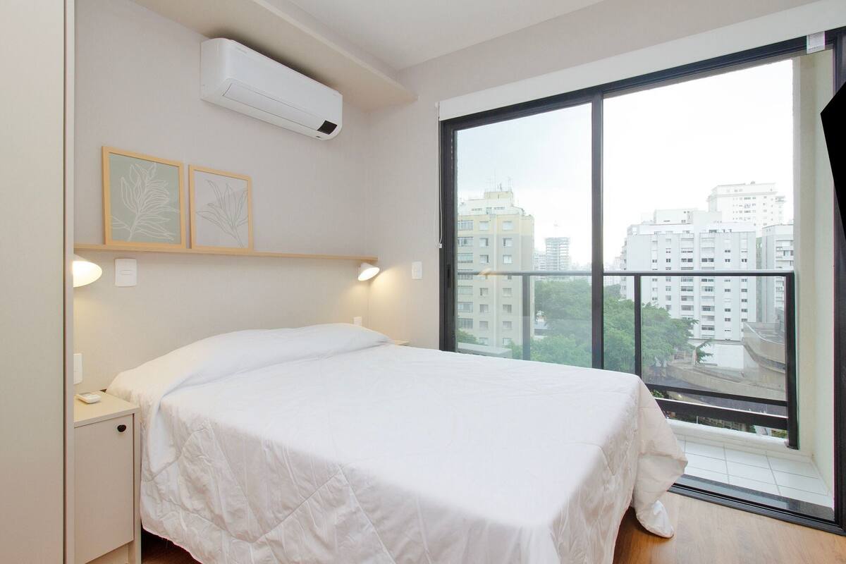 A bedroom with a bed, an air conditioner, a window with a view of buildings, and two framed pictures on the wall.