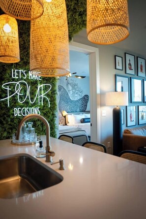 A modern kitchen with a sink, a dining table, and a wall with a green textured backdrop. There are wicker lampshades and a framed quote on the wall. - Bourbon & Broadway - Luxury Living 1.3 miles to Broadway, Brand New, 6 Beds! (Nashville)