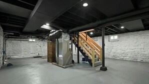 A basement with a concrete floor, a brick column, a staircase with wooden steps, and a metal duct. - FIFA Neat Nest (Raytown)