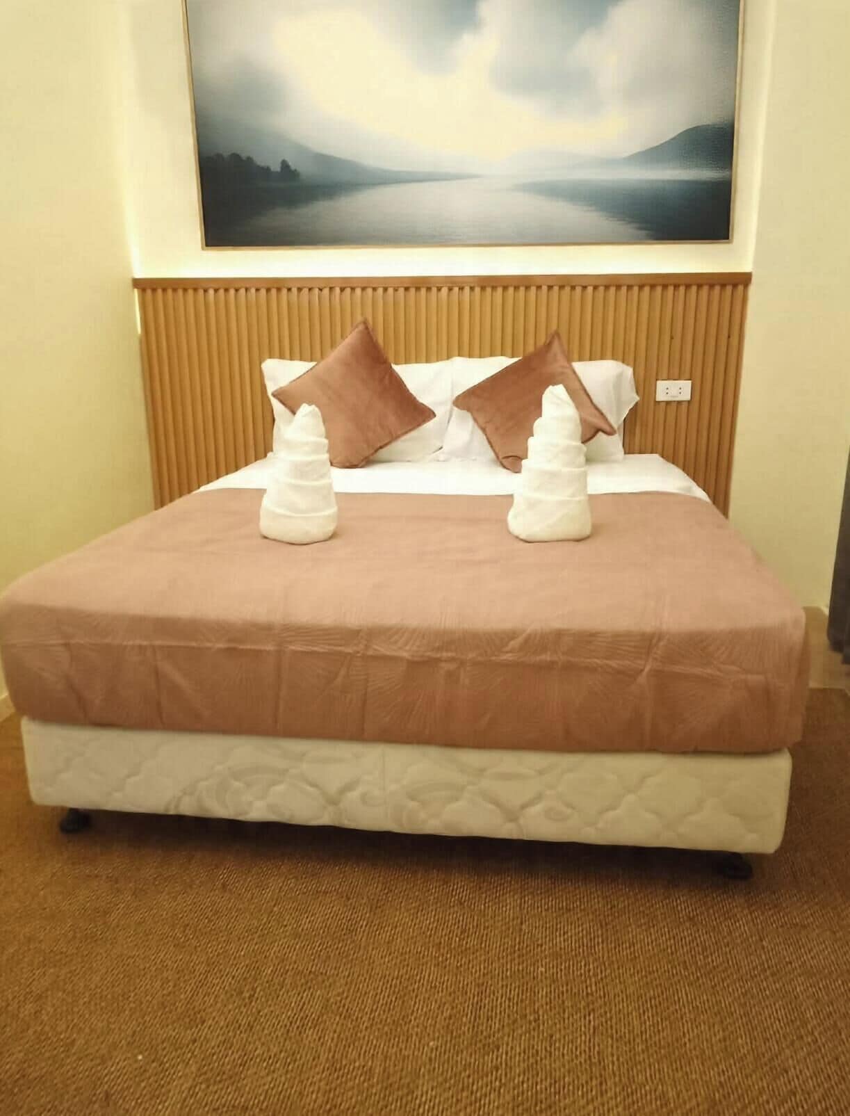 A hotel room with a bed, two pillows, a blanket, and a painting on the wall.